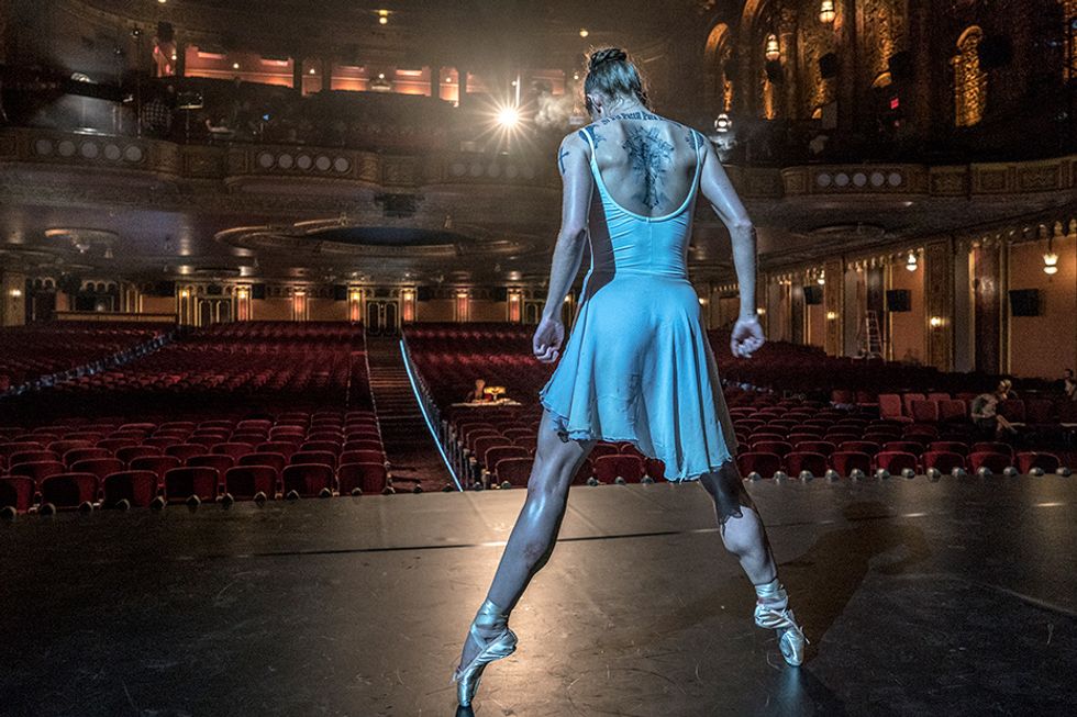 A thin white woman in pointe shoes and a white leotard and skirt ensemble balances en pointe in second position. Her arms are at her side, elbows pulled back, and her head is dropped to look at the floor in front of her. Dark, intricate tattoos are visible across her back, above the edge of the scoop-back leotard. Beyond the stage, the red seats of an opulent theater are empty, save for a figure seated at a table midway in the orchestra.